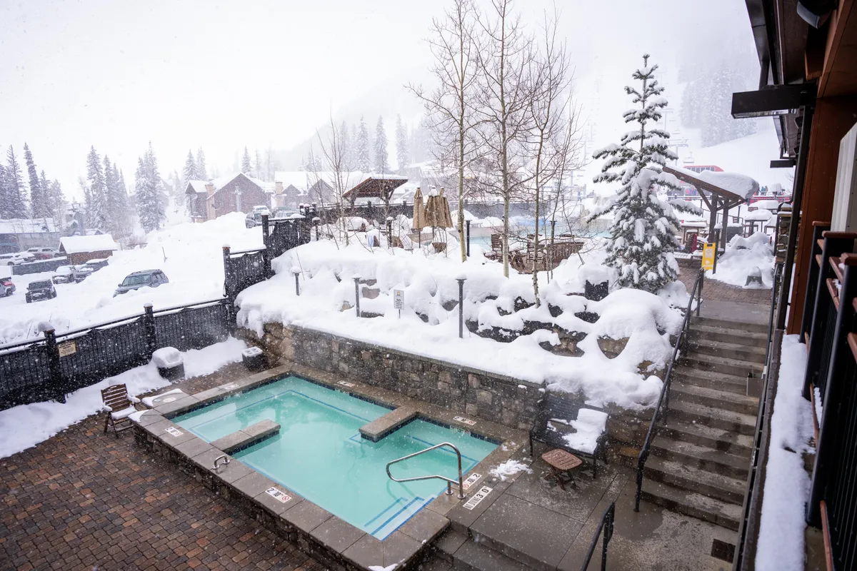 A group of people relaxes in a hot tub surrounded by snow-covered pine trees, with a staircase leading down to the steaming water. The outdoor winter environment combines nature with rejuvenation, encapsulating post-ski relaxation and social enjoyment.