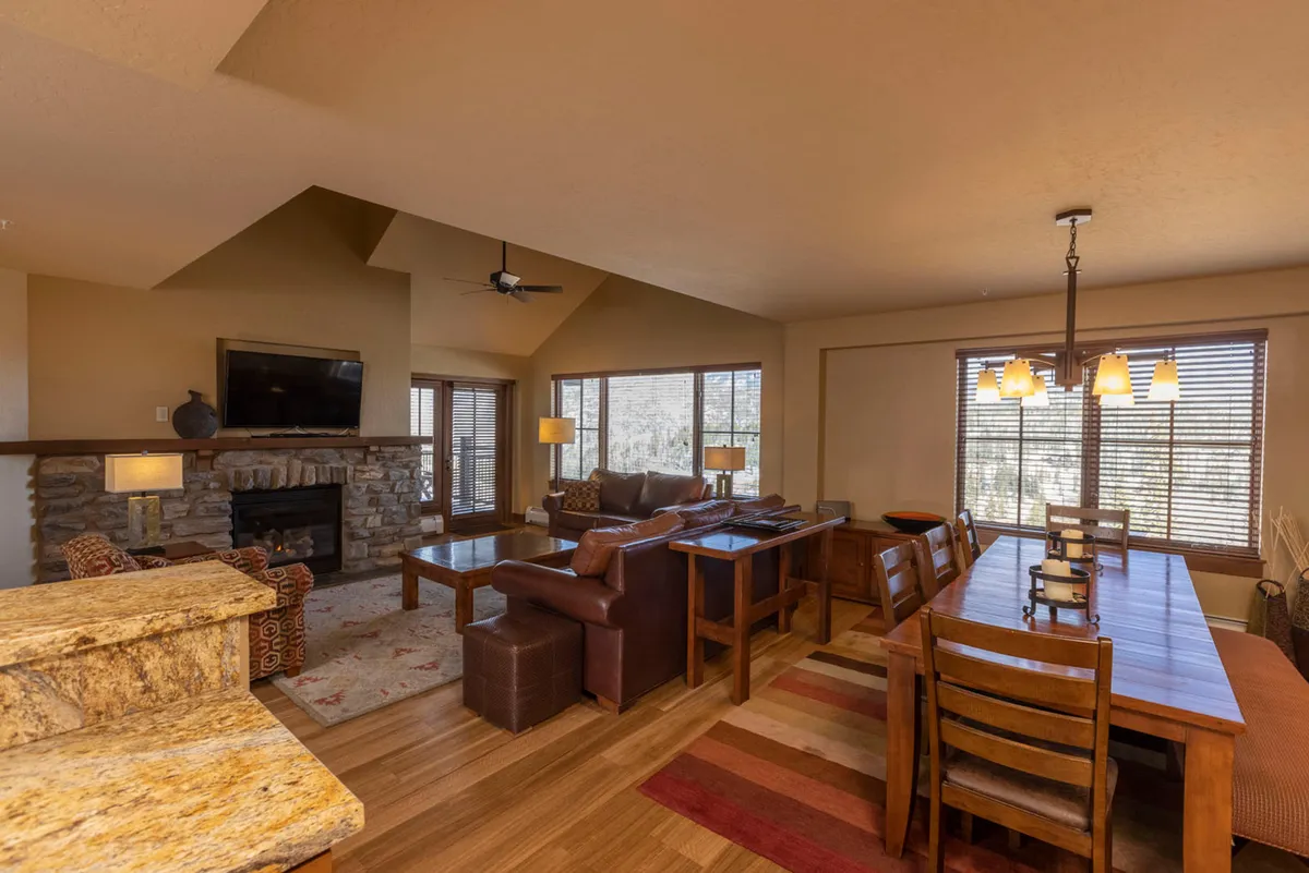 The living and dining area shown here centers around a stylish dining table set beneath vaulted ceilings, adjacent to a welcoming fireplace and ample windows. The plush seating and arrangement encourage gatherings and relaxation, with mountain views visible in the background.