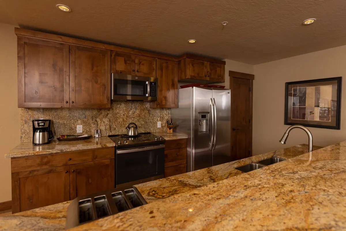 A cozy kitchen environment with sleek stainless steel appliances, expansive granite countertops, and warm wooden cabinetry. The layout is clearly designed for both practicality (ample prep space and storage) and gathering, with a coffee maker suggesting morning routines and social moments.