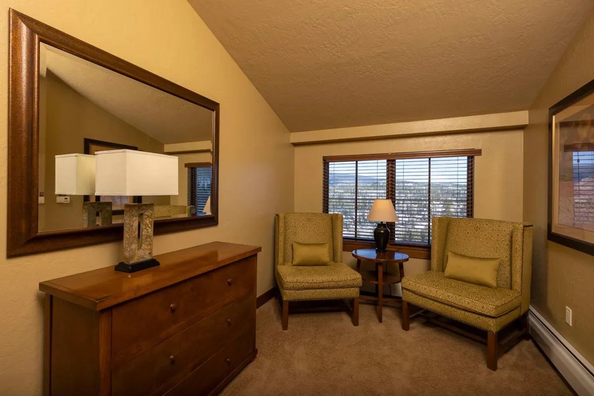 This image features two comfortable armchairs positioned near a window, a dressing table with a lamp, and a mirror. The scene is cozy and inviting, with natural light streaming in from the window, suggesting a social and relaxing atmosphere perfect for conversation or unwinding after a day of adventure.