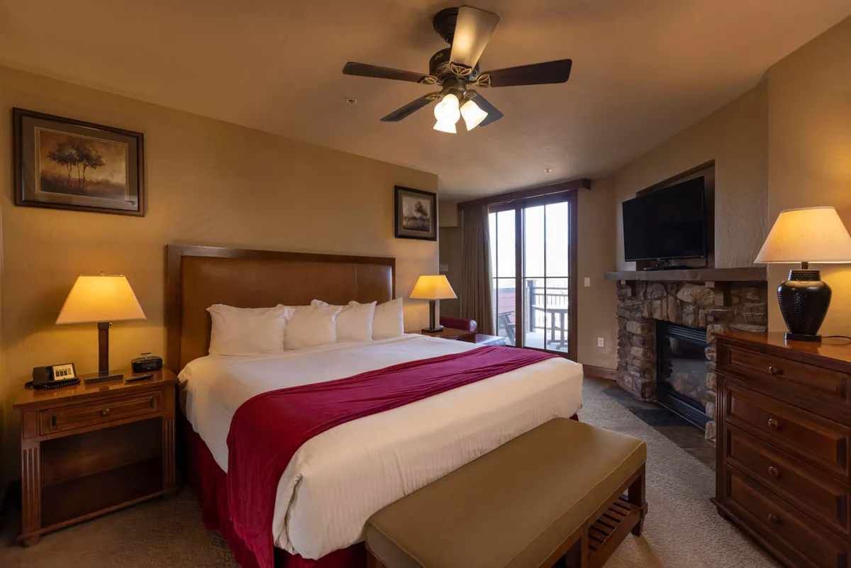 A spacious king suite features a king-sized bed with a vibrant red accent blanket, a cozy fireplace for chilly evenings, a large window letting in ample natural light, and classic wooden furnishings. The overall feel is luxurious and inviting, with relaxation zones for reading, watching TV, or simply soaking in the warmth of the fire.
