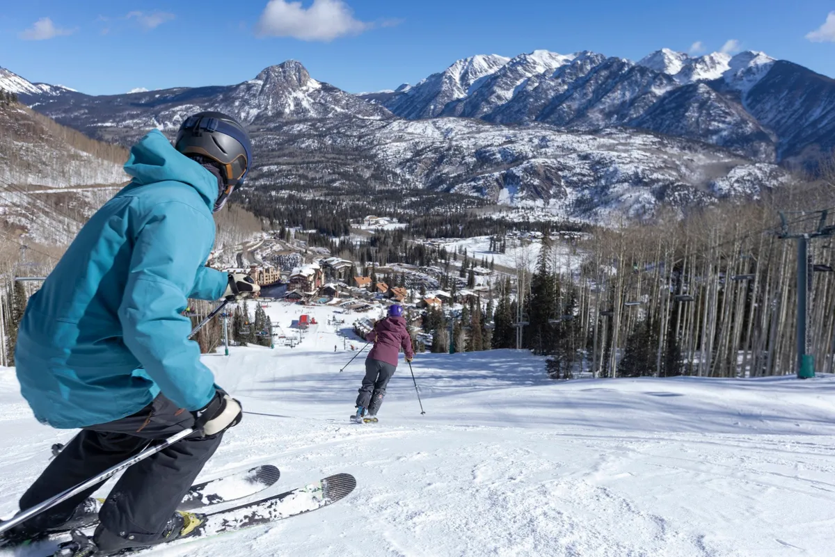 A dynamic exterior scene featuring skiers in action on pristine slopes, backed by a breathtaking mountain range and the lively atmosphere of the ski resort. The image captures both the thrill of the descent and the vibrant resort setting.