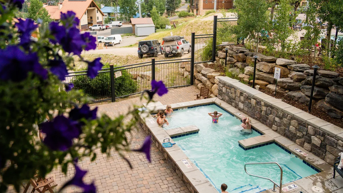 An inviting outdoor swimming pool scene, surrounded by colorful flowers and with people of all ages enjoying the water. Some guests are relaxing in the hot tub while others are socializing poolside, all beneath the warm glow of the evening sun.