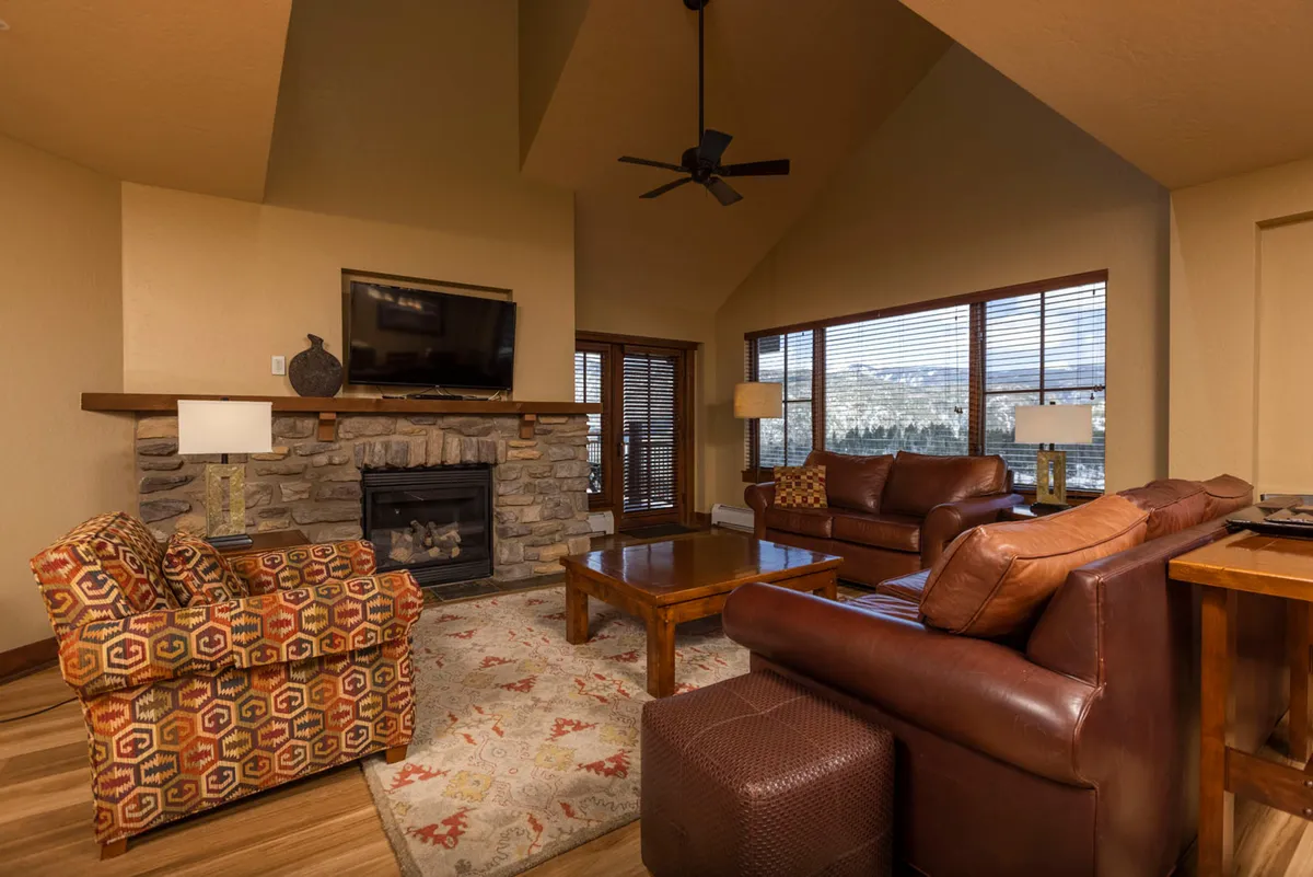 A warmly lit living area featuring a prominent stone fireplace, plush seating, and expansive windows that frame panoramic views of the surrounding landscape. Natural light pours in, enhancing the inviting atmosphere and drawing attention to both mountain vistas and the cozy, well-appointed interior.