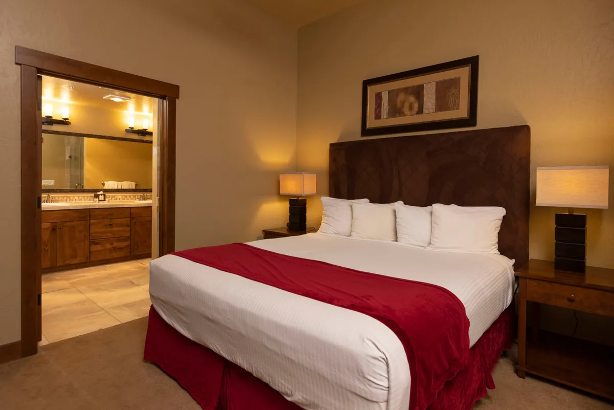 A queen bedroom with a bed dressed in red accents, flanked by modern nightstands and lamps, and a clear view of an ensuite or nearby bathroom entrance. The ambiance is restful and bright, perfect for unwinding after a busy day.