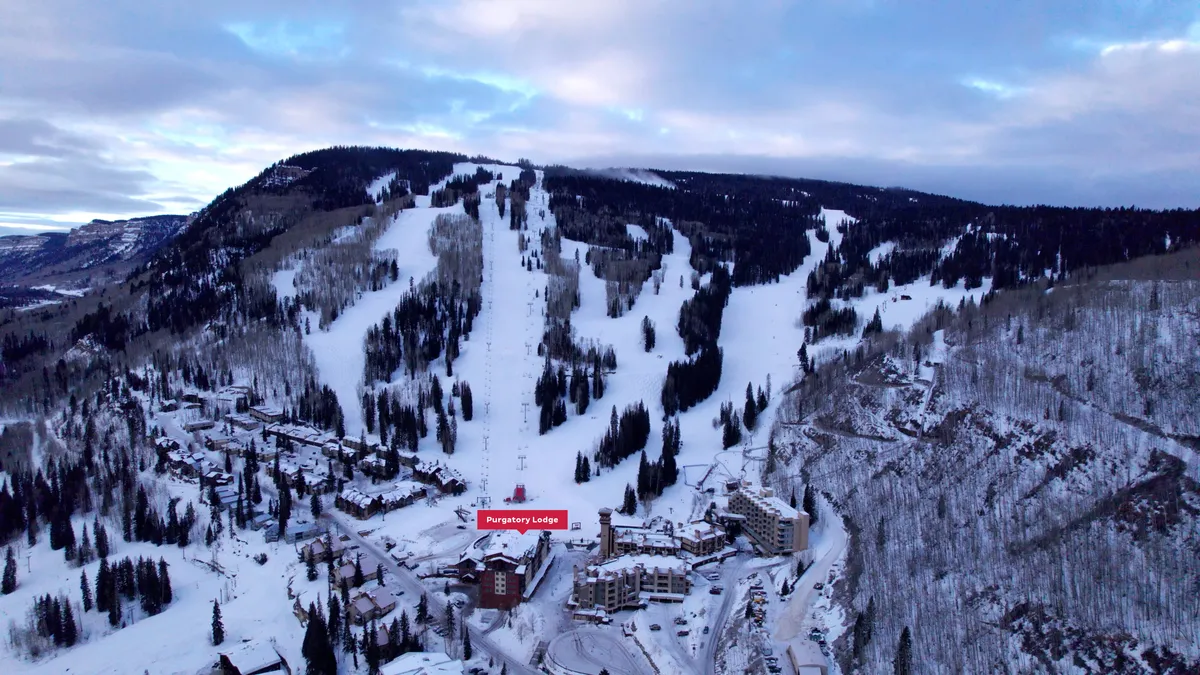 A sweeping exterior shot of Purgatory Lodge, nestled at the base of snow-covered ski slopes, with surrounding trees and a majestic mountain landscape. Ski runs and snowboard tracks are visible, emphasizing the lodge's proximity to the lifts and direct slope access.