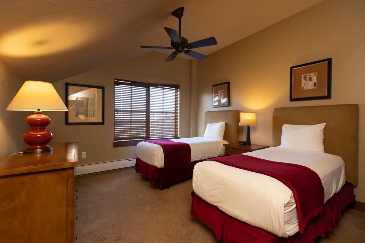A bright and cheerful twin bedroom with two beds adorned with red accent pillows, a table lamp in-between, and soft natural light streaming in through a window. The space is perfect for children or family members, with simple yet inviting decor, and a sense of coziness and calm.