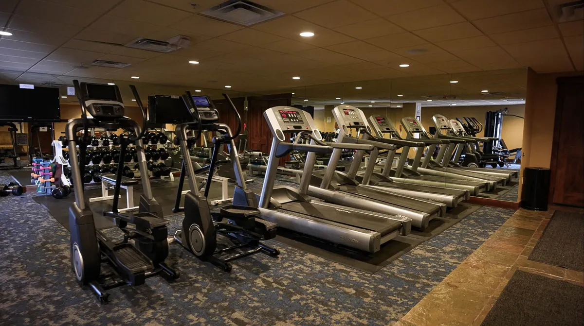 An expansive fitness center equipped with elliptical machines, treadmills, dumbbells, and a mirrored wall, set in a bright, motivating environment. The space looks ready for vigorous workouts or quiet personal training, framed by an inviting, modern design.
