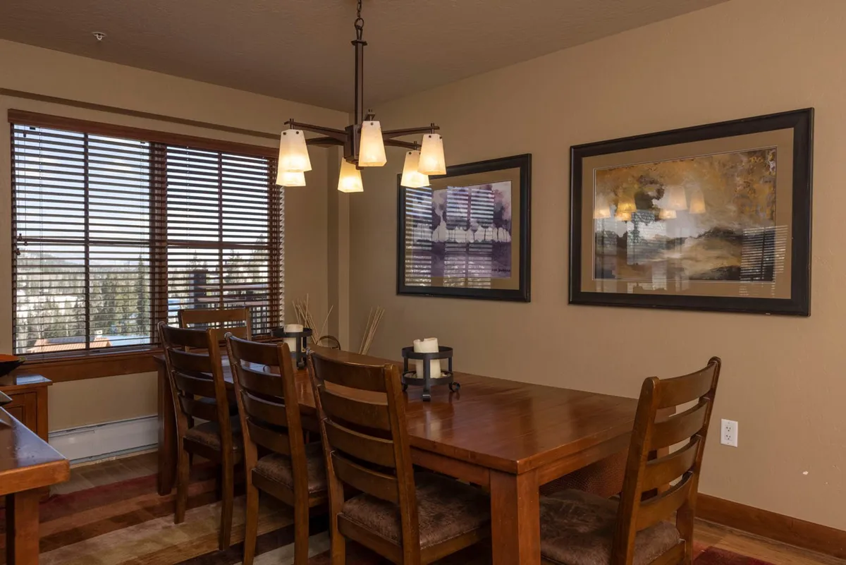 This image features a stylish dining table under a sparkling chandelier, with artwork on the walls and a window offering scenic views. The setup invites family dinners, game nights, or festive gatherings and shows how the dining space seamlessly connects to the kitchen area.